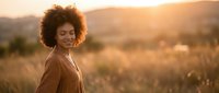 Young African American woman with curly afro hair smiling joyfully in golden sunset light outdoors in natural field landscape for lifestyle and wellness content.