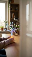 Hands holding glowing tree with colorful light branches on wooden desk in bright home office with bookshelf and plants, representing growth and innovation for business concepts.