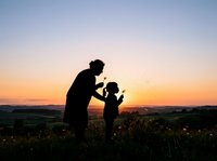 Silhouetted adult and child playing together in wildflower field at sunset, capturing joyful outdoor family bonding moments during golden hour.