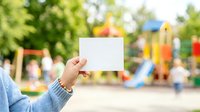 Child's hand holding blank white card at outdoor playground with colorful play equipment and blurred children in background, suitable for advertising, messaging, and promotional campaigns.