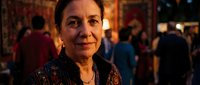 Middle-aged Hispanic woman smiling warmly at camera in elegant restaurant setting with soft lighting and blurred diners in background for hospitality concepts.