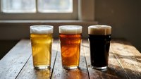Three different beer glasses with light lager, amber ale and dark stout on rustic wooden table by window. Craft brewery tasting flight comparison.