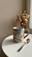 Handmade ceramic mug with marbled blue and brown glaze on white round table with dried flowers bouquet and silver spoon in warm natural light interior.