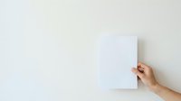 Caucasian hand holding blank white paper sheet against neutral wall background, suitable for mockup and branding projects.