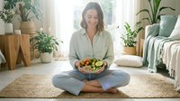 Woman in casual light green shirt and jeans sitting cross-legged on woven rug holding bowl of fresh salad in bright plant-filled bedroom. Healthy eating, wellness lifestyle.