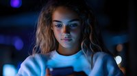 Young mixed race woman looking at smartphone screen in dark with blue light illuminating face, curly hair and white sweater, modern technology lifestyle.