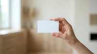 Male hand holding blank white business card against blurred interior background, suitable for branding, corporate identity, and marketing presentations.