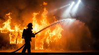 Firefighter battles intense house fire with water hose as flames engulf building structure during emergency response operation at night scene.