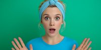 Surprised young woman with blue headband and matching shirt expressing shock against vibrant green background.