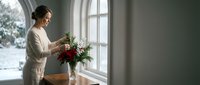 Young woman arranging fresh red roses in glass vase by window in bright modern home interior during winter season for holiday decoration and lifestyle.