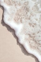 Gentle ocean foam washing onto beige sand beach creates natural diagonal composition with soft white bubbles and waves for tranquil coastal backdrop.
