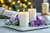 White burning candles on ceramic tray with purple lily flowers and silver Christmas ornaments against blurred bokeh lights background, creating festive holiday atmosphere.
