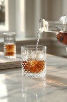 Pouring amber whiskey from crystal decanter into cut glass tumbler on marble counter with warm natural lighting creating elegant drinking atmosphere.
