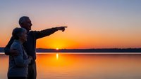 Senior couple enjoying romantic sunset by lake with man pointing toward horizon during golden hour vacation moment.