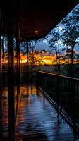 Wet wooden deck with metal railing reflects golden sunset through silhouetted trees, creating dramatic sky view with orange and purple clouds after rain.