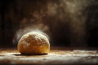 Fresh bread dough ball dusted with flour on wooden surface with atmospheric flour particles illuminated by dramatic side lighting.