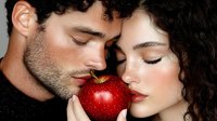 Sensual couple sharing red apple, faces close together with eyes closed against dark background. Romantic and tempting concept for relationship stories.