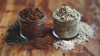 Two glass jars containing brown cocoa and light clay powder on wooden surface, natural cosmetic ingredients spilled around containers for spa and beauty treatments.