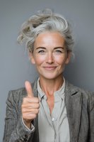 Confident mature woman with silver hair showing thumbs up gesture against gray background, wearing professional attire.