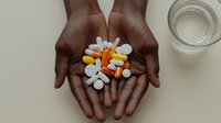 African American hands holding colorful prescription pills and capsules with glass of water nearby for medication adherence concept.