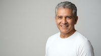 Confident mature Hispanic man with gray hair smiling warmly in white casual shirt against neutral background for lifestyle and wellness content.