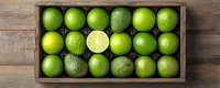 Fresh green limes arranged in wooden box on rustic table, top view. Whole citrus fruits with one cut lime showing juicy flesh, food background.