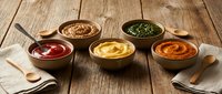 Five colorful condiment sauces in wooden bowls arranged on rustic wood table with spoons for cooking and food preparation.