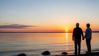 Romantic couple silhouettes holding hands watching golden sunset over calm lake water with peaceful sky reflection for love and relationship concepts.