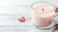 Burning pink scented candle in glass jar with decorative red paper hearts on white wooden background, creating romantic atmosphere for Valentine's day celebration.