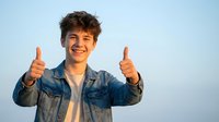 Young cheerful man with curly hair giving double thumbs up gesture wearing denim jacket against clear sky background expressing positivity and success.
