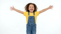 Happy mixed-race child with curly hair in yellow shirt and denim overalls celebrating with arms raised wide, expressing joy and excitement on white background.