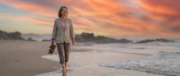 Woman in casual beige clothing walking barefoot along sandy beach at sunset, carrying shoes, ocean waves and dramatic sky in background.