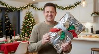 Happy middle-aged Caucasian man in beige sweater holding wrapped Christmas gifts in festively decorated home with tree and garland during holiday season.