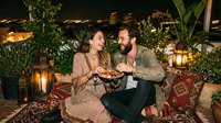 Young Caucasian couple sharing food on a boho rooftop terrace at night, surrounded by lanterns, lush plants, and colorful rugs.
