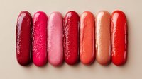 Row of glossy lipstick swatches in various shades of red and pink on beige background, showcasing color range for makeup application.