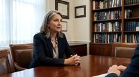 Middle-aged woman with long gray hair and fair complexion, wearing professional dark blazer and striped blouse, seated at wooden conference table in office library setting during business meeting or interview.