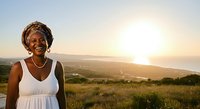 Senior African woman in white sleeveless dress smiling at sunset over rolling hills and grassland landscape during golden hour.
