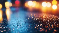 Colorful bokeh lights reflecting on wet asphalt surface during rain at night, creating magical atmosphere with golden and blue blurred circles and water drops.