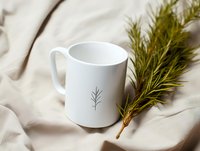 White ceramic mug with minimalist pine branch design on fabric background alongside fresh pine twigs, creating cozy winter atmosphere for seasonal marketing.