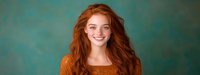 Young woman with flowing red hair and bright smile wearing orange sweater against turquoise background, natural beauty portrait in panoramic format.