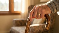 Senior man with weathered hands holding wooden cane indoors, symbolizing aging, mobility assistance, healthcare, retirement living, and elderly care support needs.
