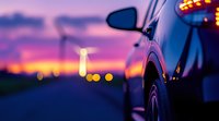 Car side mirror and body reflecting purple and orange sunset sky with blurred city lights in background, creating atmospheric evening road scene.