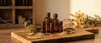 Amber glass essential oil bottles with dried herbs on rustic wooden board in warm natural light for aromatherapy and wellness products.