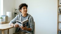 Young man of mixed ethnicity reading book in modern home office with shelving and desk, suitable for education and lifestyle content.