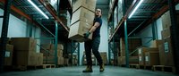 Young Caucasian male warehouse worker carrying heavy cardboard boxes in a large storage facility with shelving units and inventory.