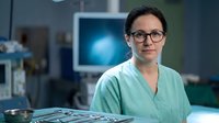 Female surgeon in green scrubs and glasses standing confidently in modern operating room with medical equipment and monitors in background.