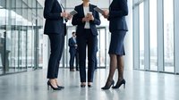 Business professionals in formal attire shake hands in a modern office lobby with glass walls, symbolizing partnership, agreement, and corporate collaboration in a contemporary workplace setting.