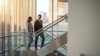 Two professional business people walking up modern glass staircase in contemporary office building with city skyline view through windows.
