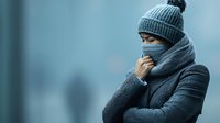 Young Asian woman wearing protective face mask, winter coat and knit hat standing outdoors on cold foggy day, expressing illness symptoms and discomfort.