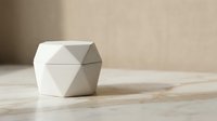 White geometric ceramic container with angular faceted design on wooden surface against neutral beige wall background for modern home decor and storage.